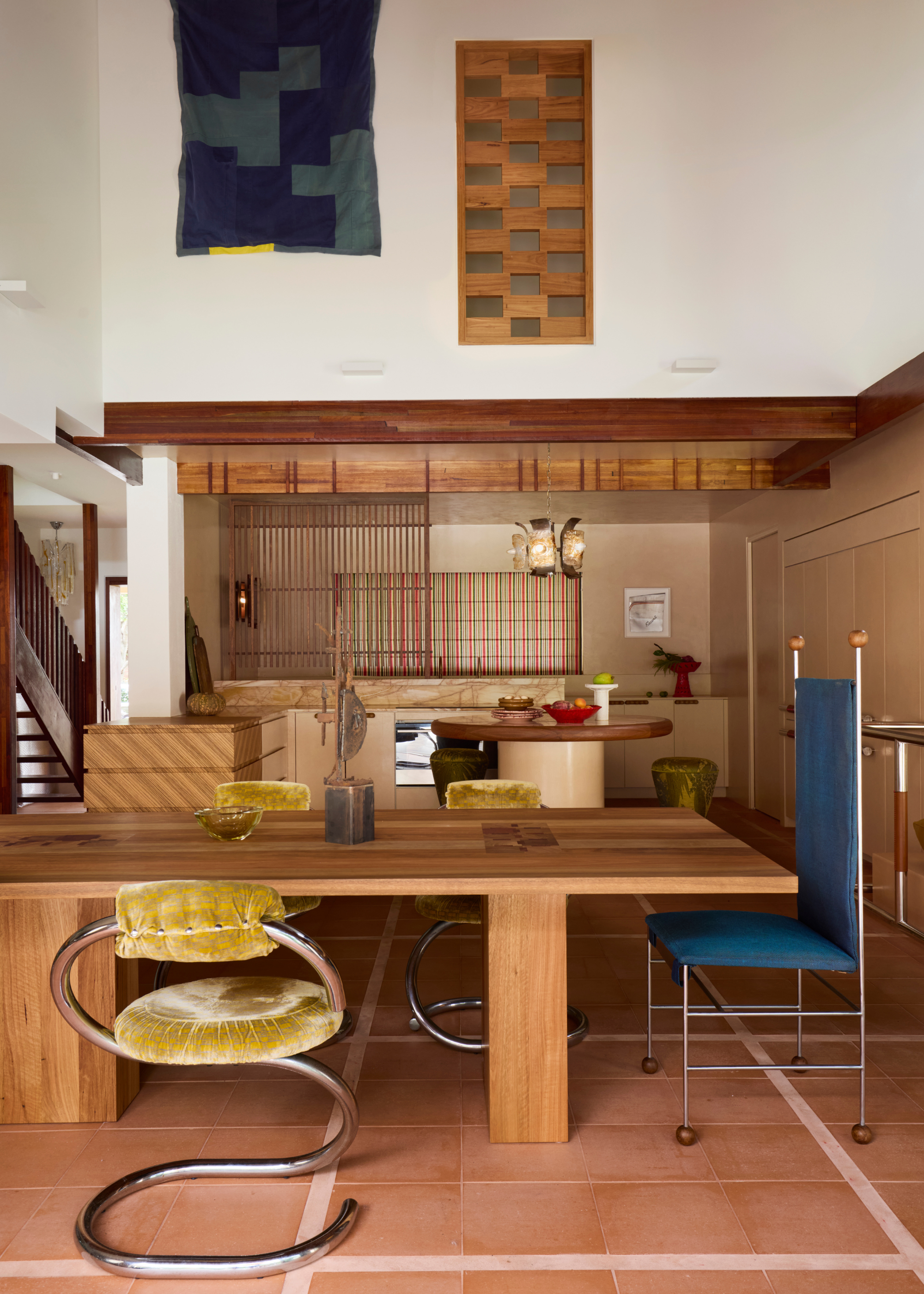 An open plan living area with high white ceilings with a handwoven wall hanging next to a timber divider, a large wooden dining table with chrome dining chairs with shimmery green backrests and seats with a circular kitchen island and kitchen area at the back of the space