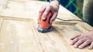 Person using an electric hand sander on bare wood panel door