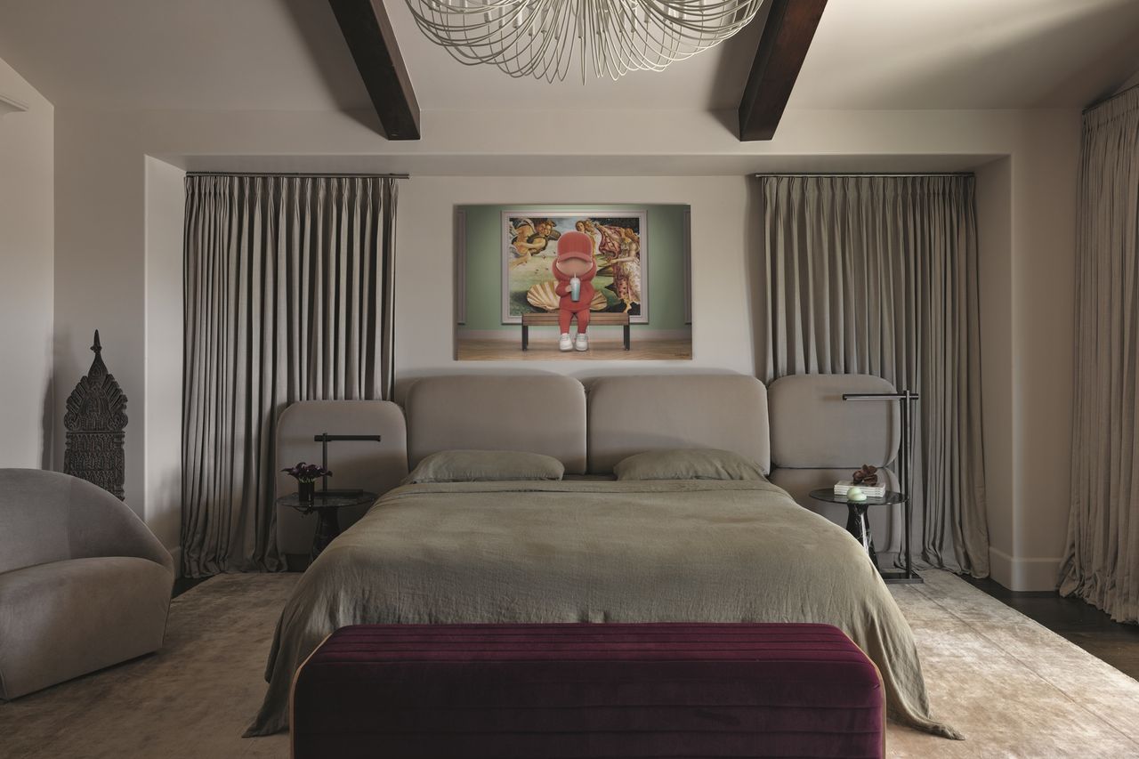 modern bedroom with beige walls, ceiling beams, modern chandelier, artwork hanging above bed, curtains on either side of the bed, burgundy ottoman at the foot of the bed, gray-green bed linen, gray bedhead, beige rug, gray sofa, and heavy drapes