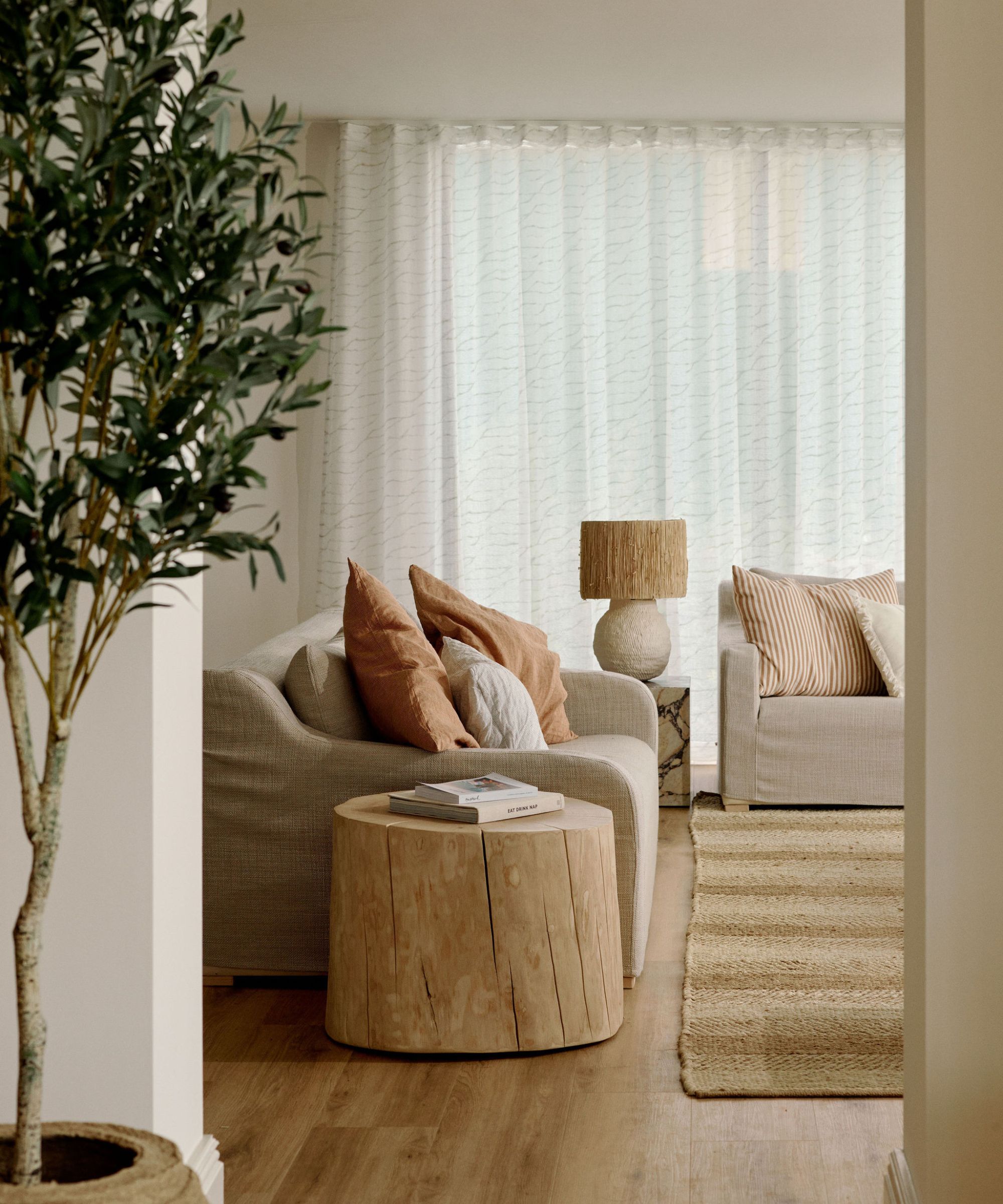 neutral warm minimalist living room with a jute rug, two linen slip covered sofas, a rustic wooden side table and an olive tree