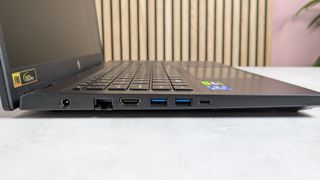 Left side ports of Acer Nitro V 15, open on desk with wooden panels, plant, and pink wall in background