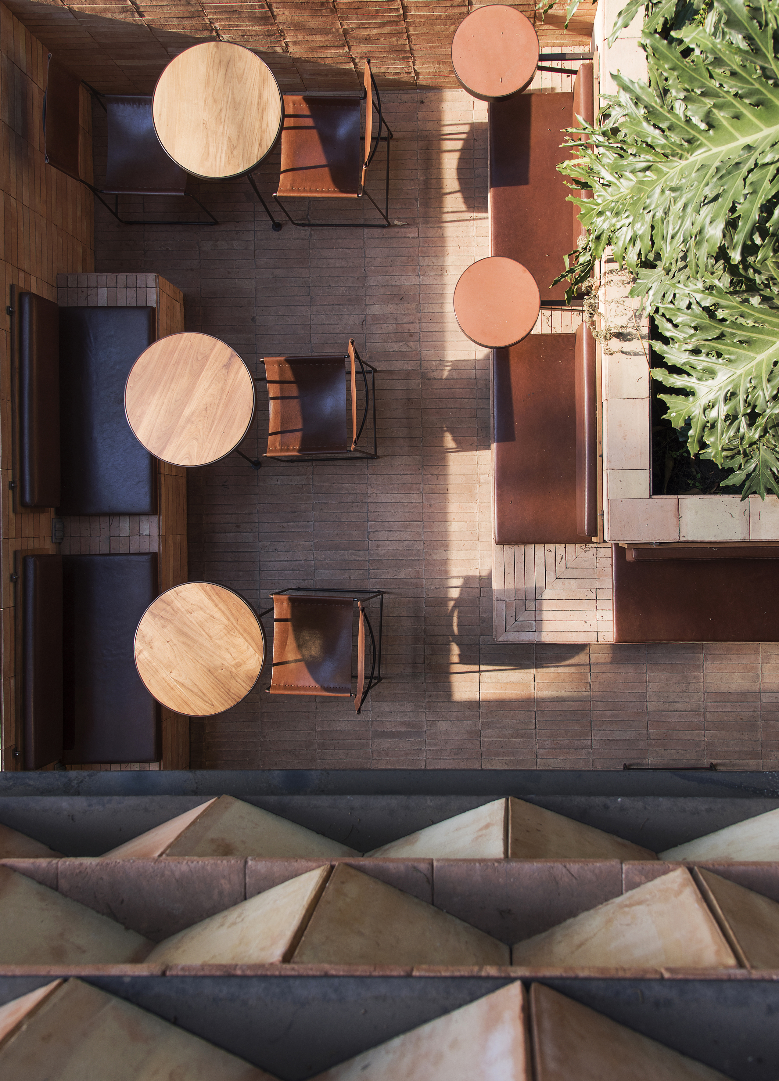 An earthy-tinted, brutalism-inspired cafe's outdoor patio features terracotta-clad surfaces and geometrically shaped, minimalist furnishings mixing in with verdant vegetation.