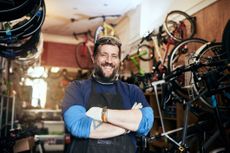 A burly man who own a bicycle shop smiles while standing on the shop floor.