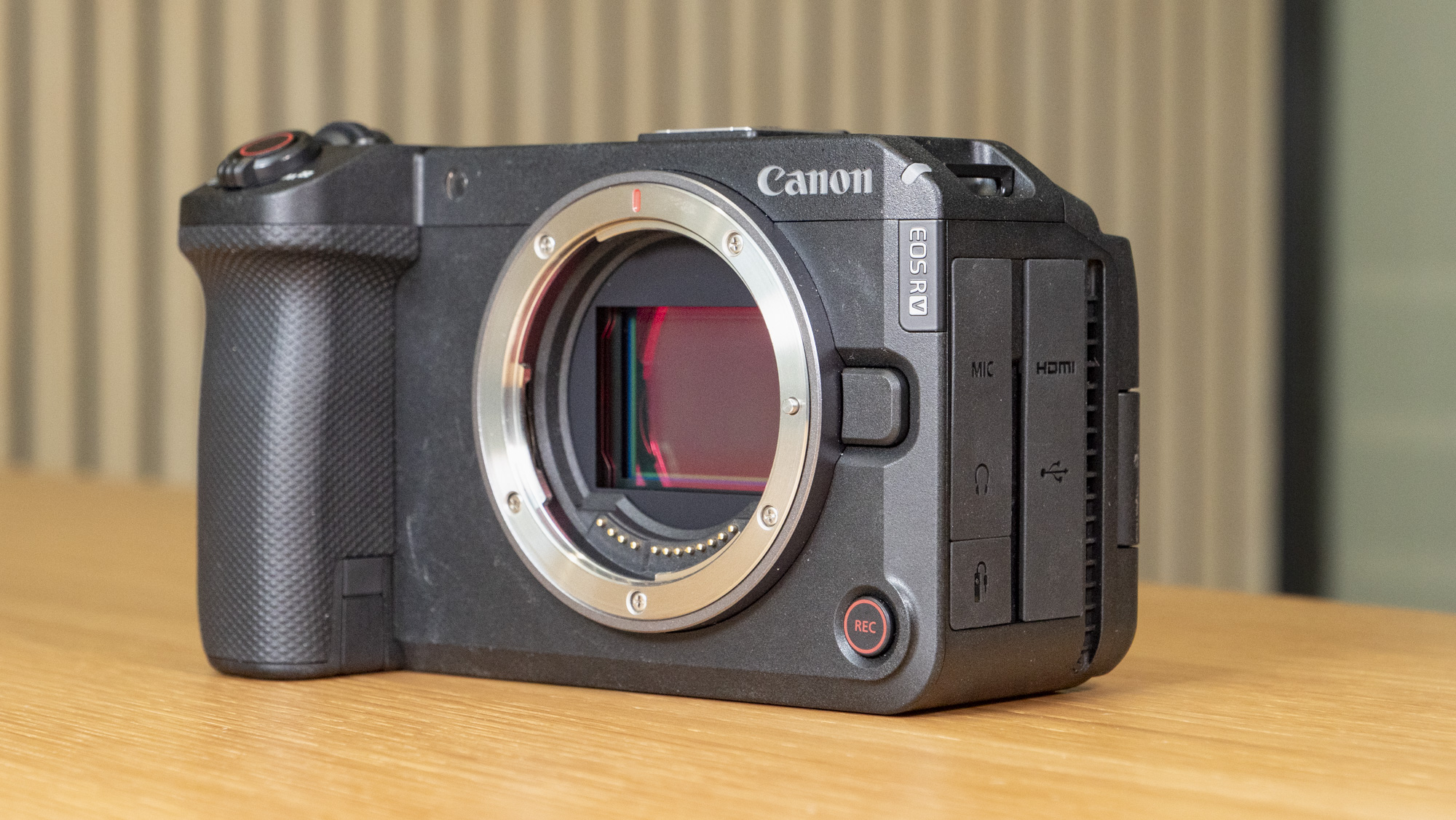 The Canon EOS R6 V mirrorless camera on a wooden table with a pine-slatted backdrop, no lens attached
