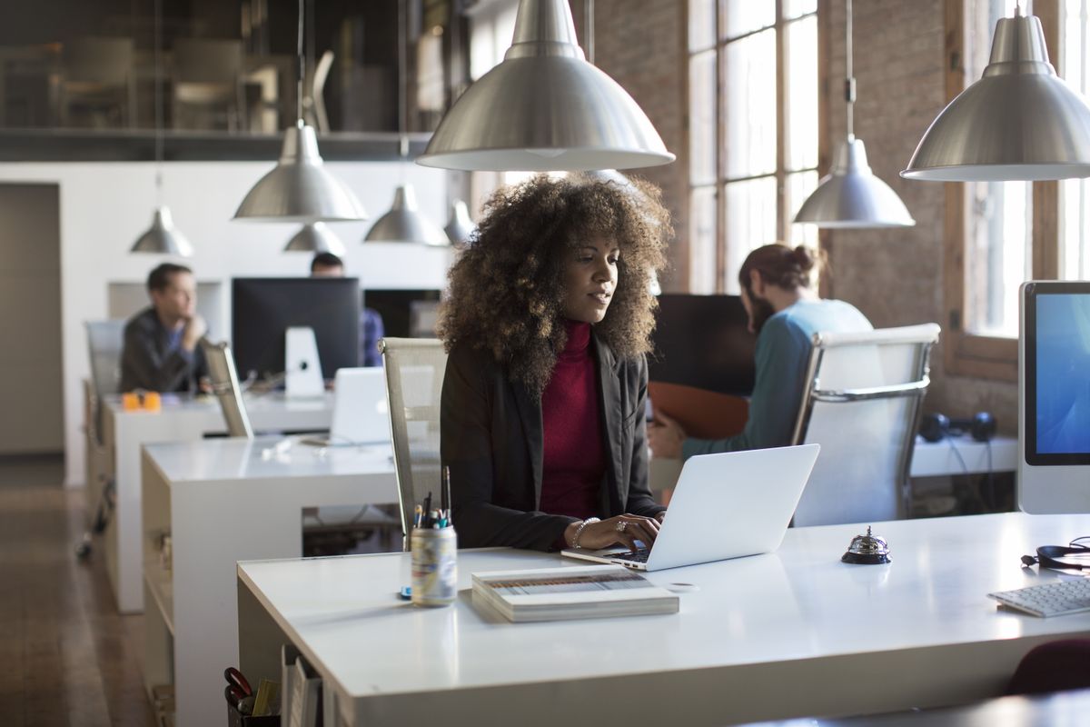 A woman sitting alone at her desk in a tech startup office space with men in the background