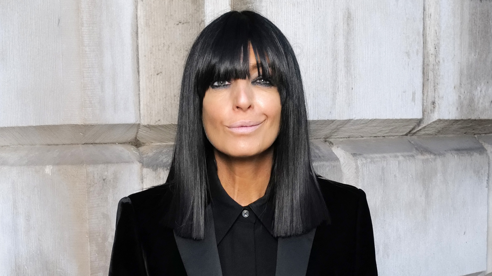 Image of Claudia Winkleman with black straight bob hair with fringe, smiling, wearing a black shirt and blazer