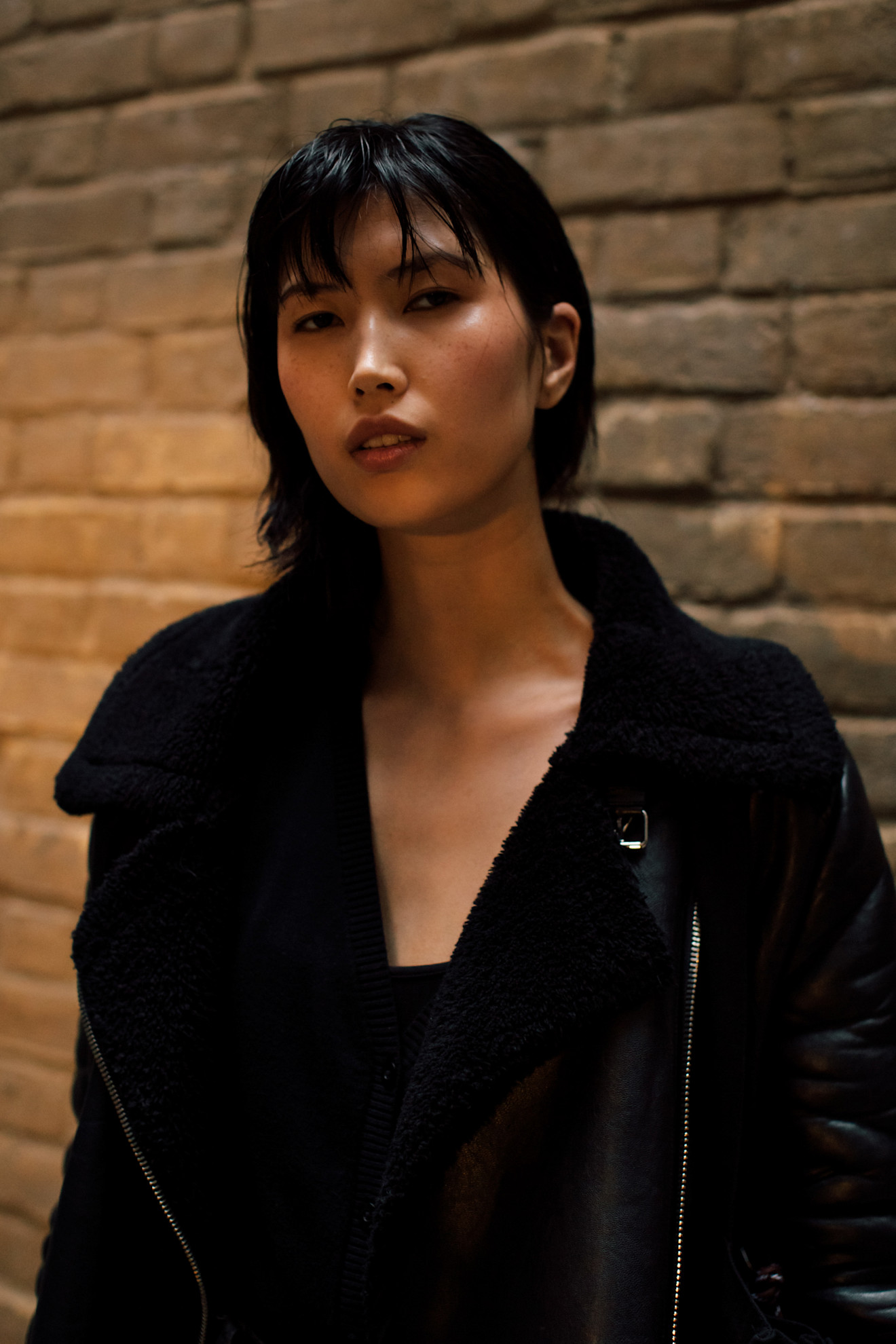 a woman in a black jacket with black hair in a bob against a brick wall