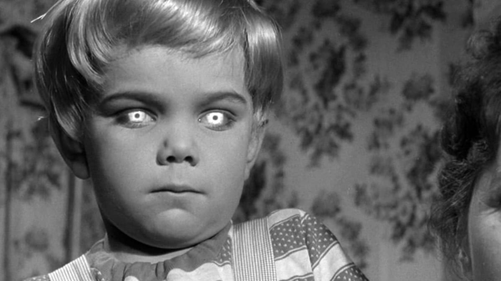 A child with blonde hair has shining/evil-looking eyes and looks straight back at the viewer