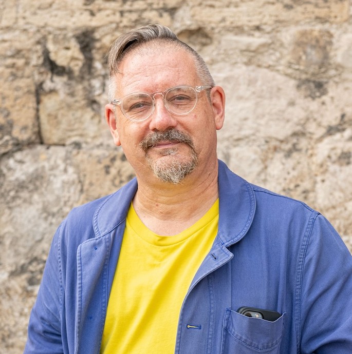 A man with glasses and a grey beard wearing a yellow T-shirt and a blue jacket