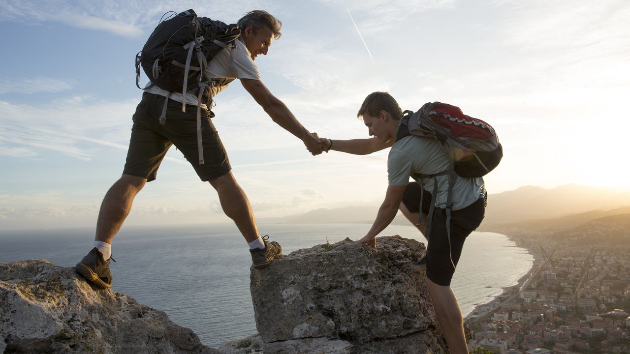 A father gives his adult son a hand up on a mountain.