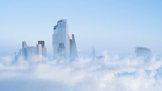 An aerial view of the London Financial District, with clouds superimposed over the buildings to represent finance in the cloud. The buildings are set against a hazy blue sky.