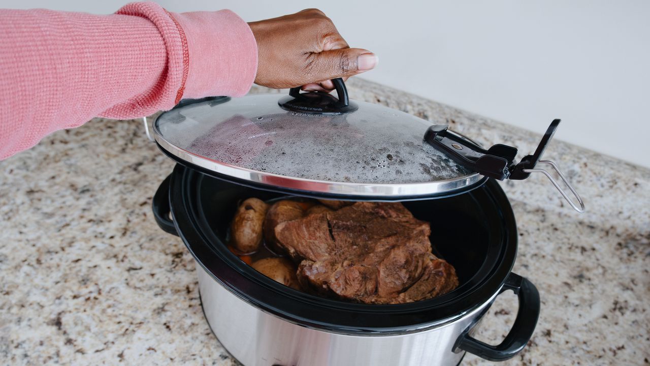 Hand lifts lid on slow cooker with chuck roast and potatoes visible