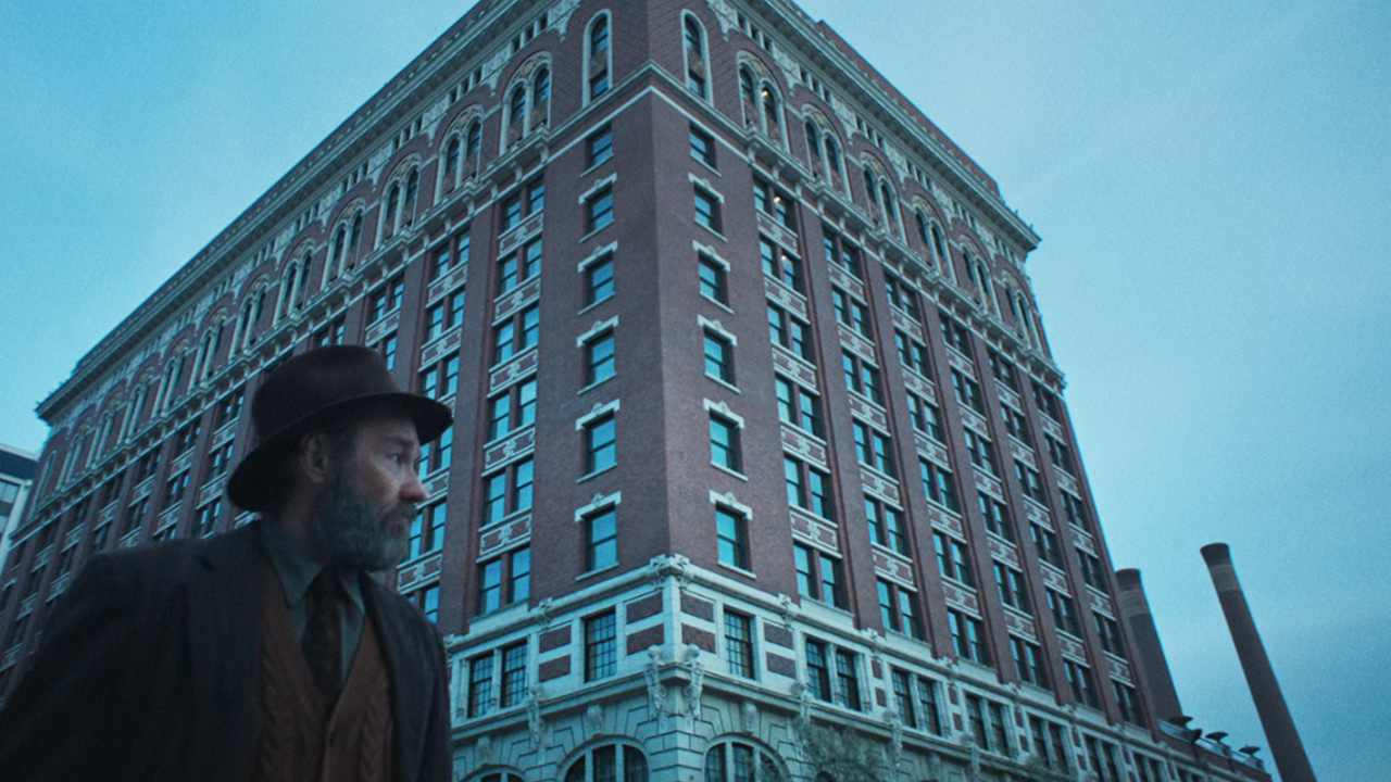 Joel Edgerton in a hat and suit, walking in front of a tall building