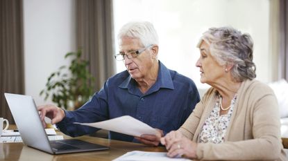 An older couple look over paperwork while sitting at a table together with a laptop.