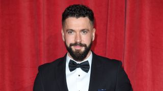 Shayne Ward wearing a black suit and bowtie in front of a red background