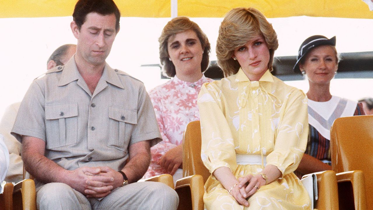 Prince Charles looking down and wearing a tan shirt sitting next to Princess Diana wearing a yellow dress and frowning