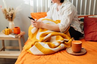 A woman sitting in her bed which has orange bedding on it, with a stripey yellow blanket and a candle.