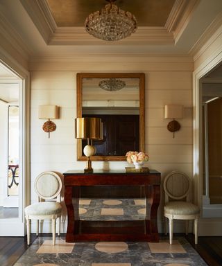 Large and lavish entryway space painted in rich beige, accented with lamps, mahogany console and layered lighting