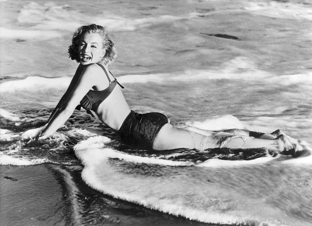 Black and white image of Marilyn Monroe lying on a beach amidst the surf