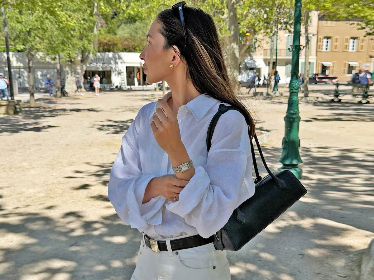 Style influencer Felicia Akerstrom in a square in Saint Tropez wearing black sunglasses on her head, a white button-down shirt, a black Alaia Le Teckel shoulder bag, black belt, and ivory jeans.