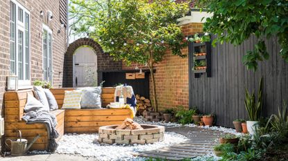 Small garden with wooden outdoor seating, fire-pit and wooden boardwalk on white gravel, black wooden fence