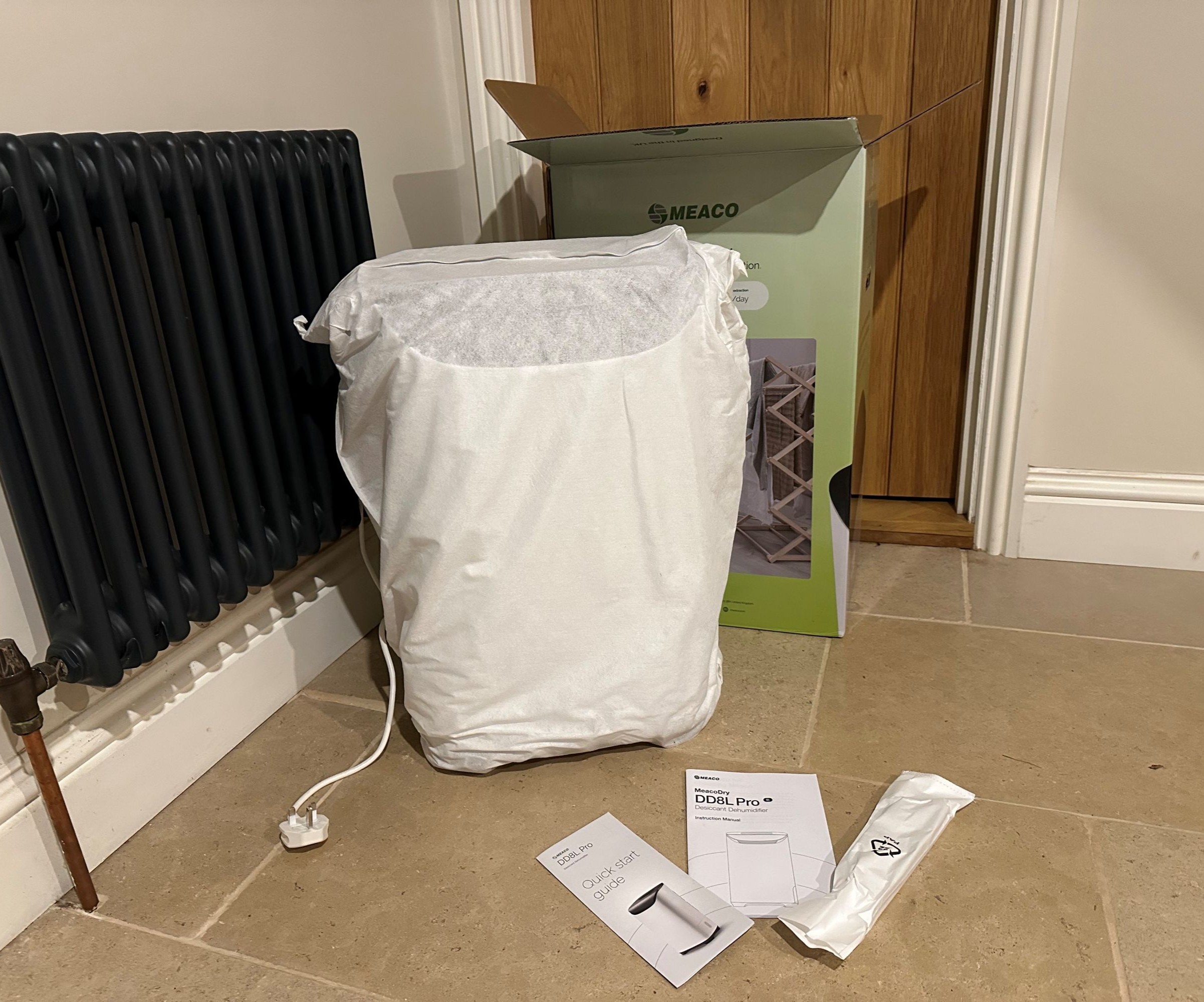 The Meaco dehumidifier during unboxing and still in its dust cover with the box behind it and the instructions laid on the stone floor in front of it