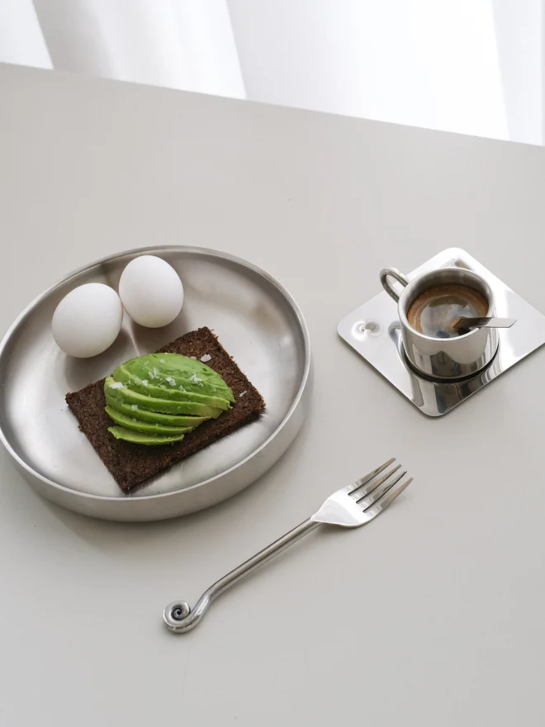 A stainless steel bowl from SOOS Atelier with avocado on rye bread and boiled eggs, beside a swirl ended fork, and a chrome coffee coaster set