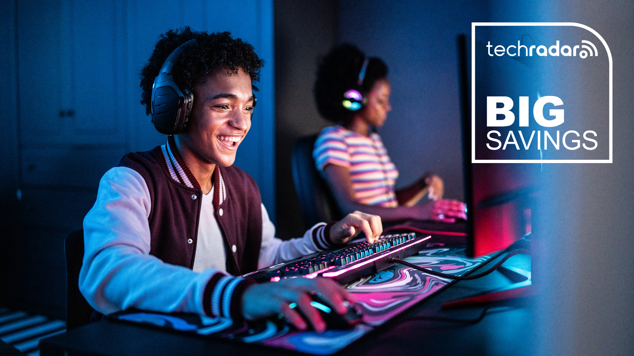 Two teenage gamers sitting side-by-side at a large desk while wearing headphones, using a mouse and keyboard and each looking at a bright monitor that illuminates their faces in a darkened room.
