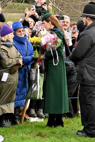 Kate Middleton wears black boots, a long green coat, a green hat, and carries flowers on Christmas Day at Sandringham