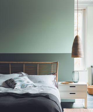 A two toned green bedroom, with a lighter color on the top half of the wall and darker color on the bottom half. a bed with a rustic wooden bed frame and low hanging rattan pendant.
