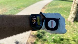 The Garmin Venu X1 on a wrist next to a golf glove, showing the yardage for the hole, with a golf cart path and tree visible behind the Venu X1.