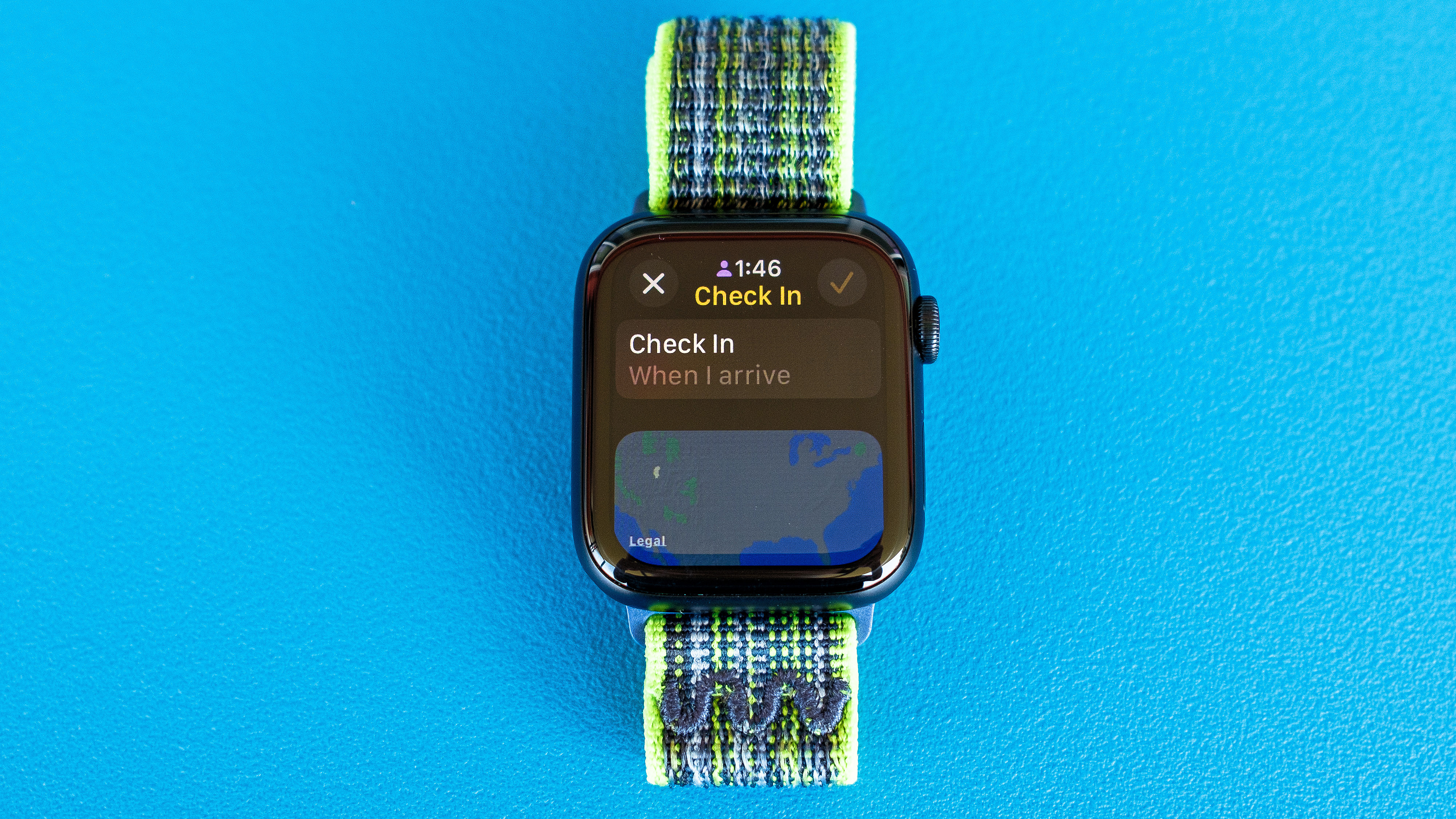 Gambar detail Apple Watch SE pada latar belakang biru cerah yang menunjukkan langkah-langkah untuk menyiapkan dan mengirim pemberitahuan Check In di watchOS 11. Gambar ini menunjukkan cara Check In melalui tujuan