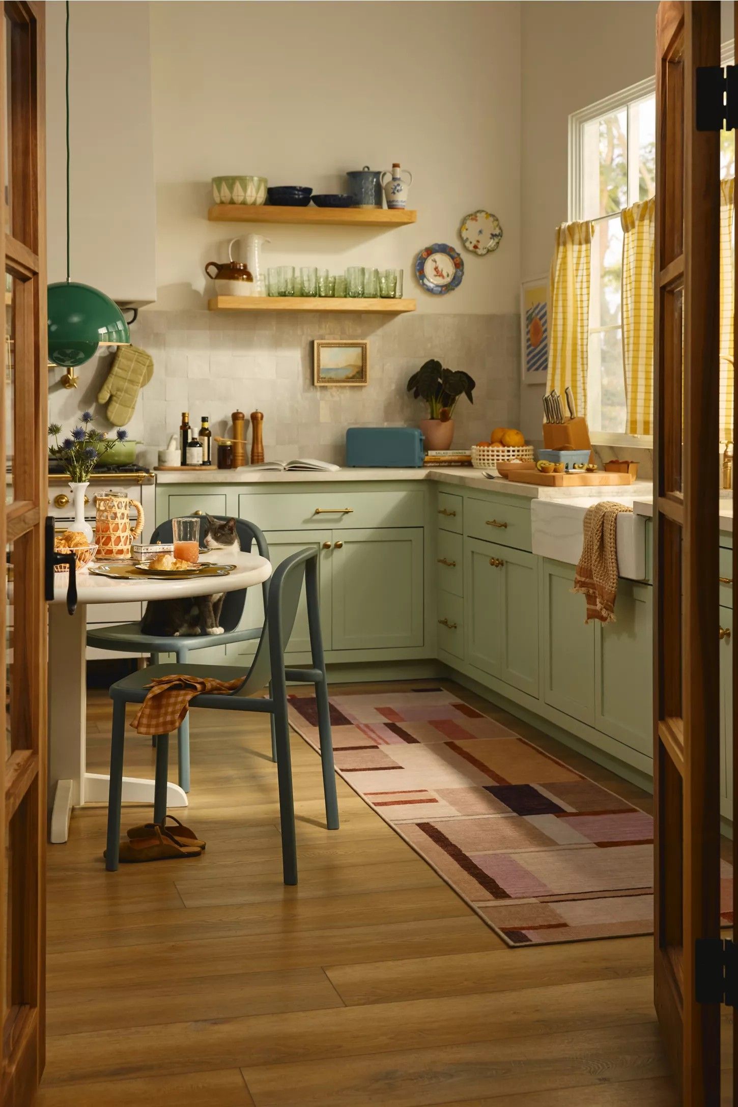 Image of a retro-inspired kitchen with white walls, wooden floors, and mint green cabinetry. There is decor all around the space and a pink block-print runner rug in front of the sink.
