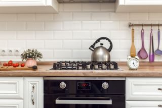 A cream kitchen with subway tile backsplash, colorful utensils on a brass bar, tomatoes on a chopping board, and steel kettle on the stove