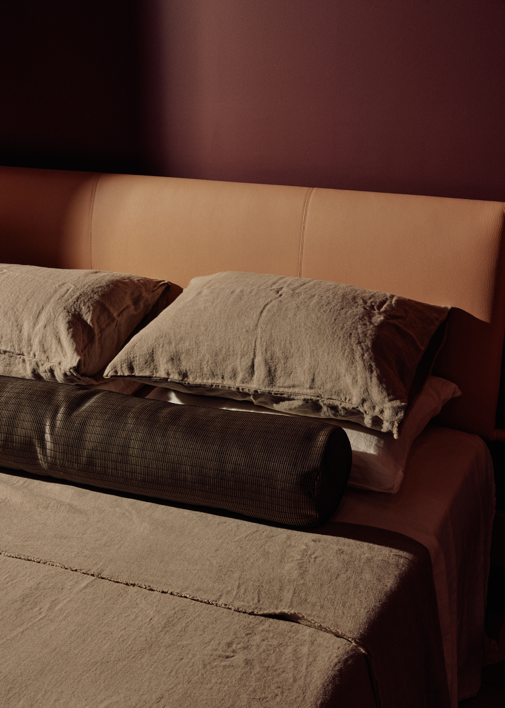 A burgundy bedroom with a tan headboard and a stack of green pillows by a green duvet and a dark bolster pillow