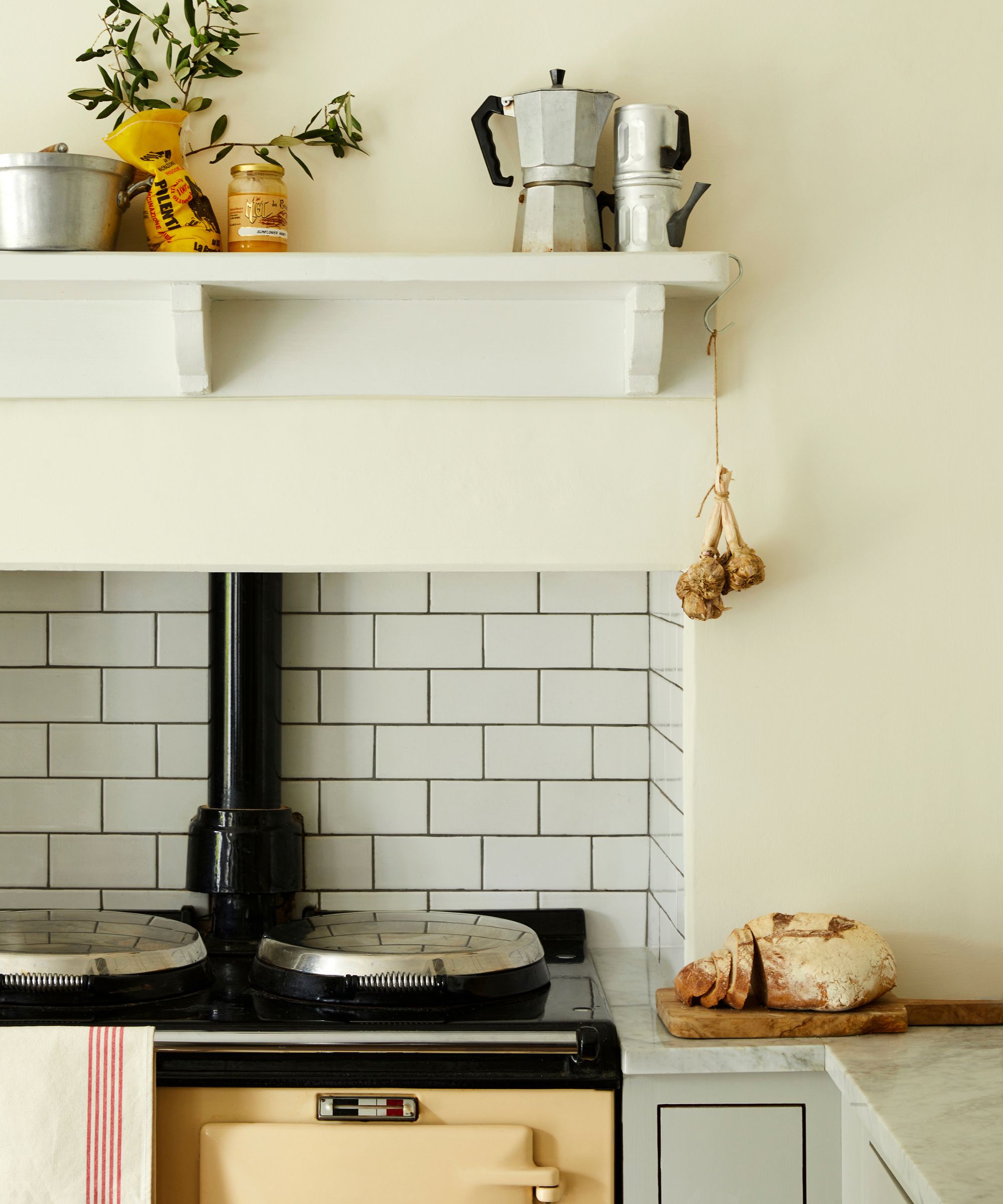 Traditional cream kitchen featuring a black range cooker with twin hotplates and white metro tile splashback. A wooden shelf above holds jars and stovetop coffee makers. Garlic hangs from a hook, and a crusty loaf rests on a marble counter.