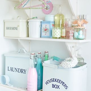 a white laundry room with white shelves, holding pastel blue laundry tin and housekeepers tin basket, along with an assortment of cleaning and laundry products
