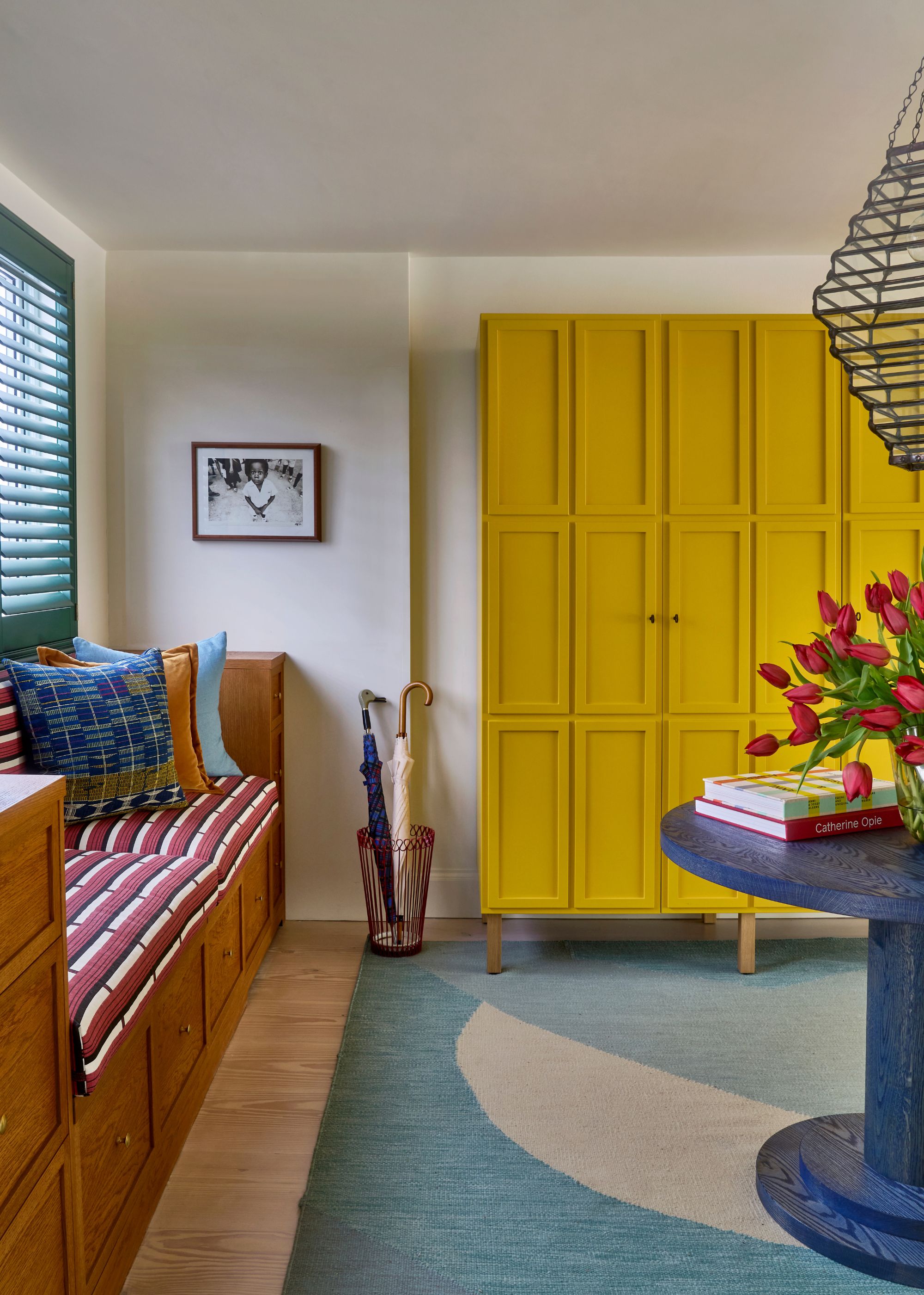 An entrance hall with white walls, bright yellow wardrobes, an aqua blue rug, a deep blue round table with a vase of red tulips, and a wooden window bench.