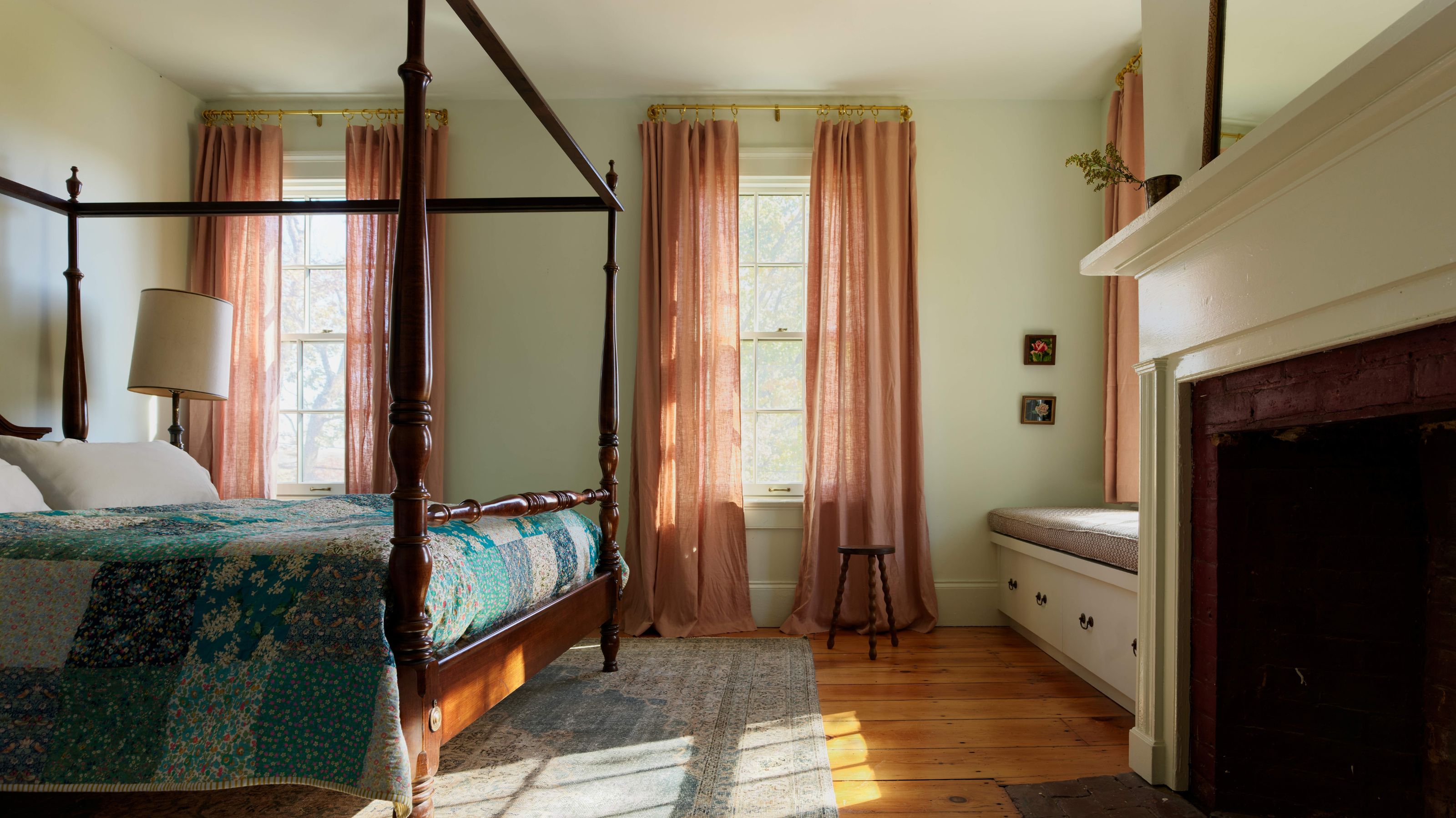 Bedroom featuring a four-poster bed with a colorful quilt, terracotta-colored curtains on two tall windows, a window seat with storage drawers, and an exposed brick fireplace