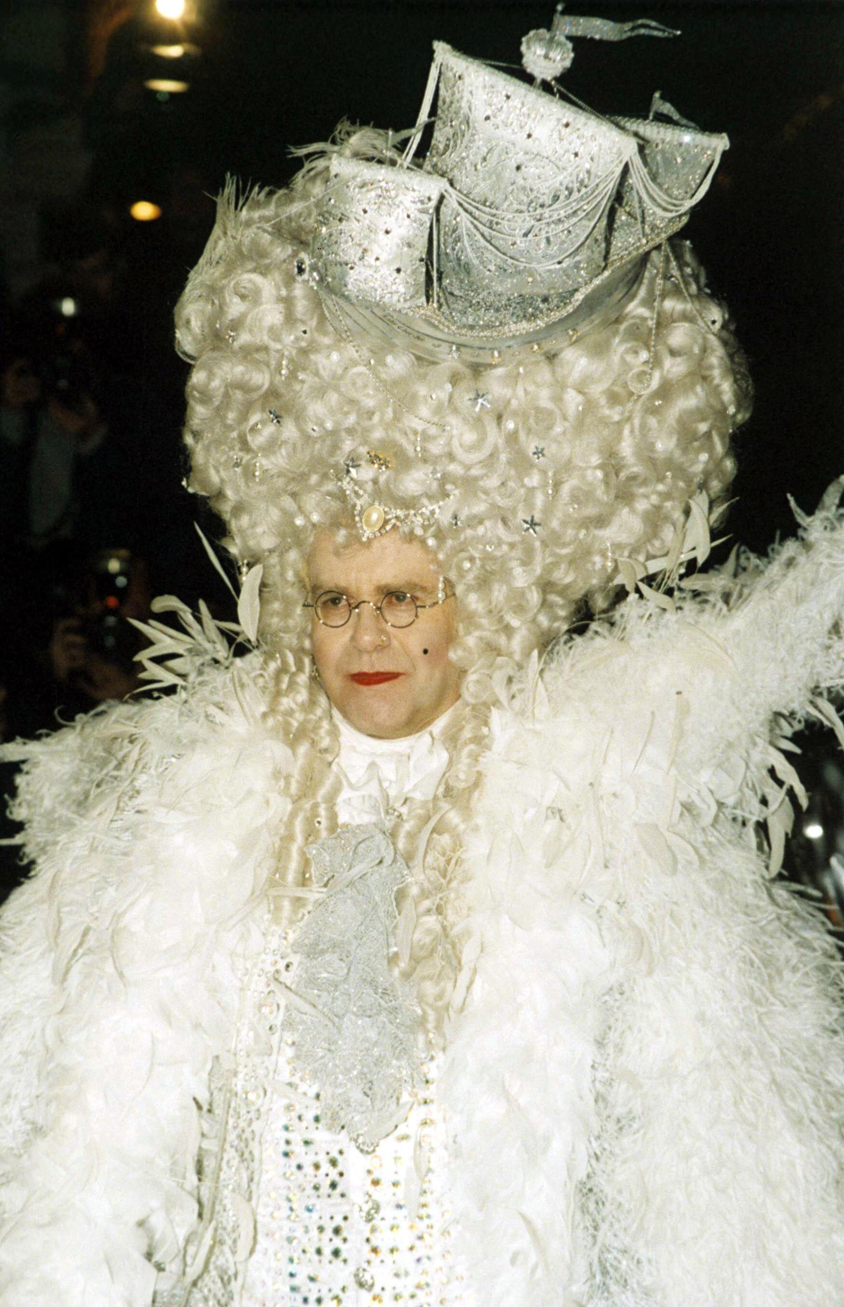 Elton John in his iconic outfit for his 50th birthday. He is wearing a white gown, a large white wig and a ship on his head