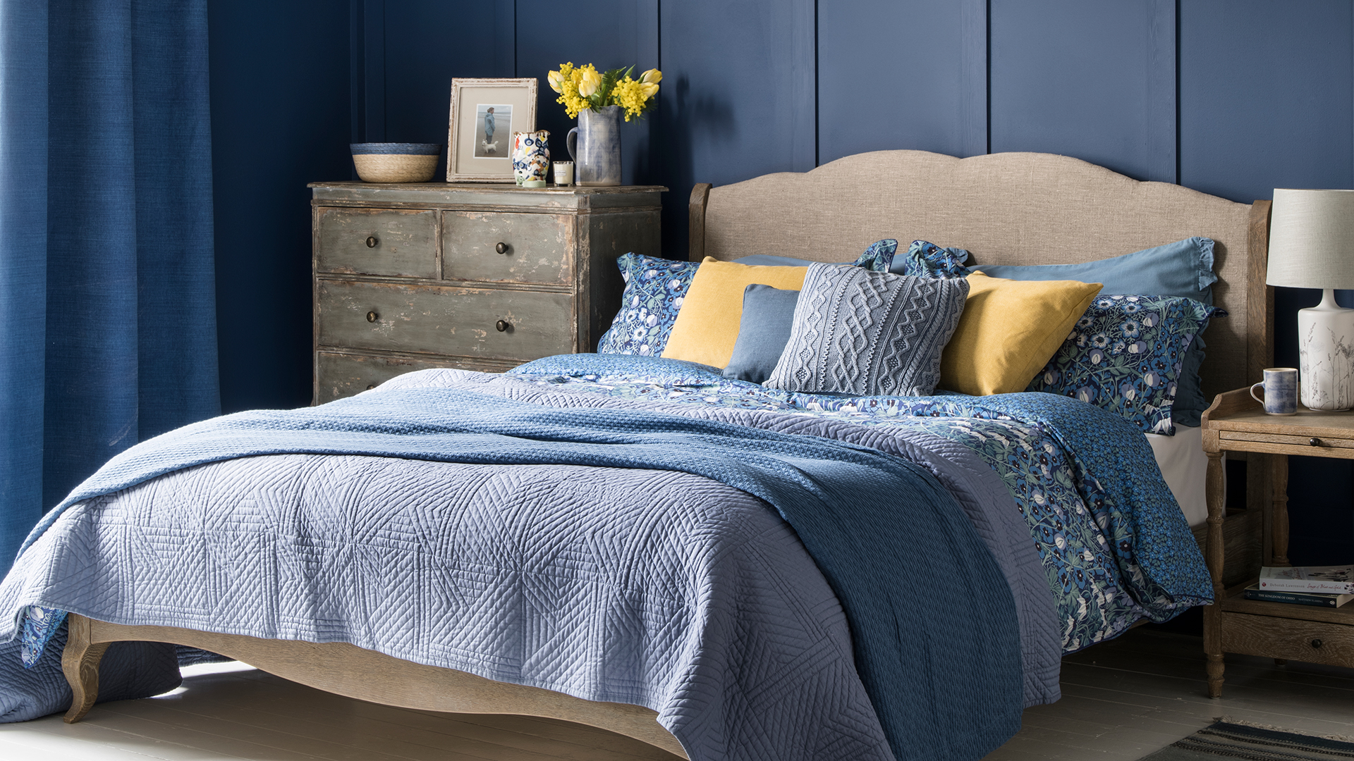 Navy blue bedroom with a neutral linen upholstered and wooden bed with blue and yellow bedding