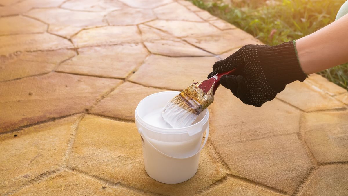 hand wearing black glove holding paintbrush above tub of white paint stood on yellow concrete patio slabs