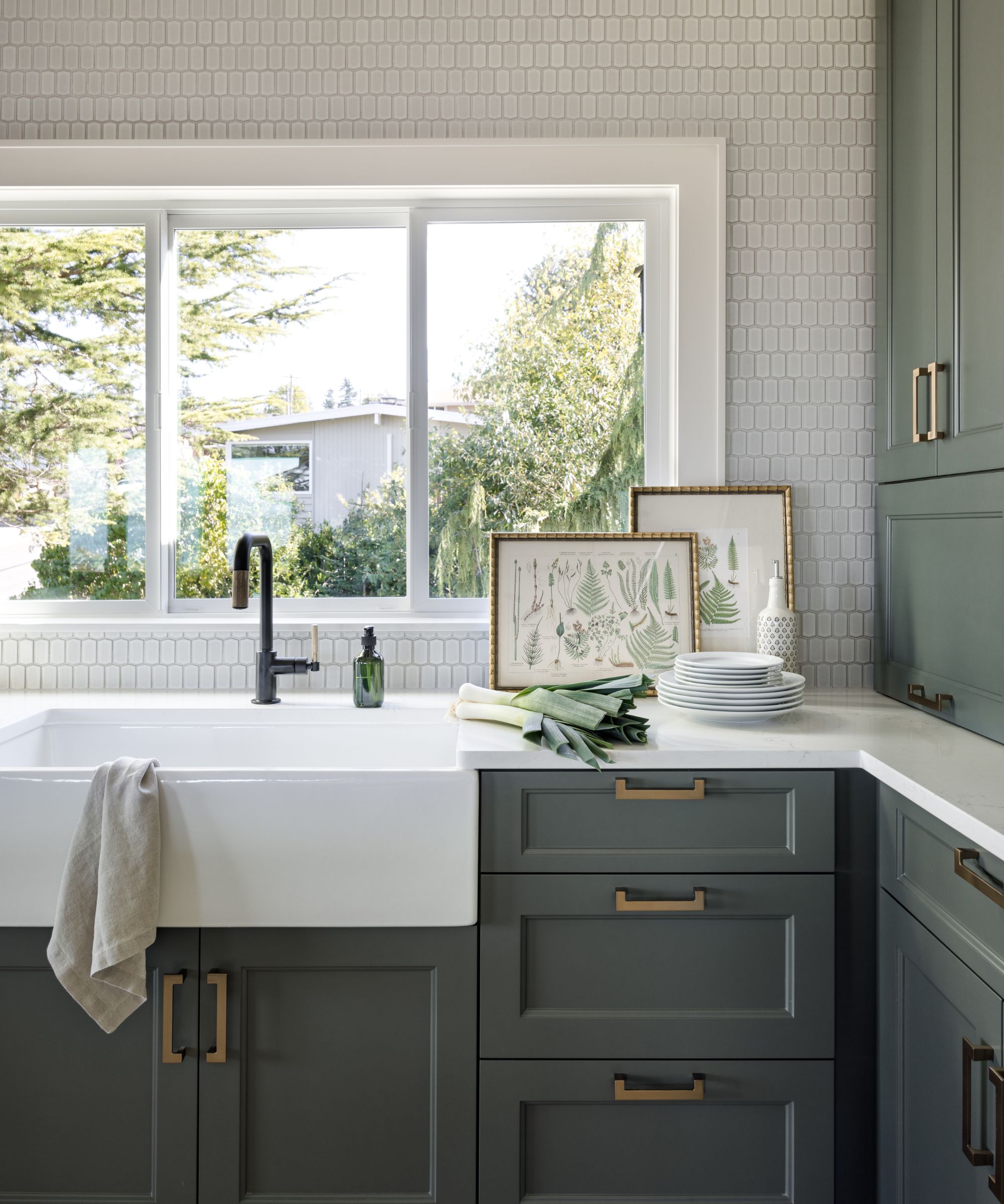 The corner of a small kitchen with dark gray-green cabinets with brass handles, a white Belfast sink in front of a window with a black tap. White countertops and two framed pictures leaning against the window.