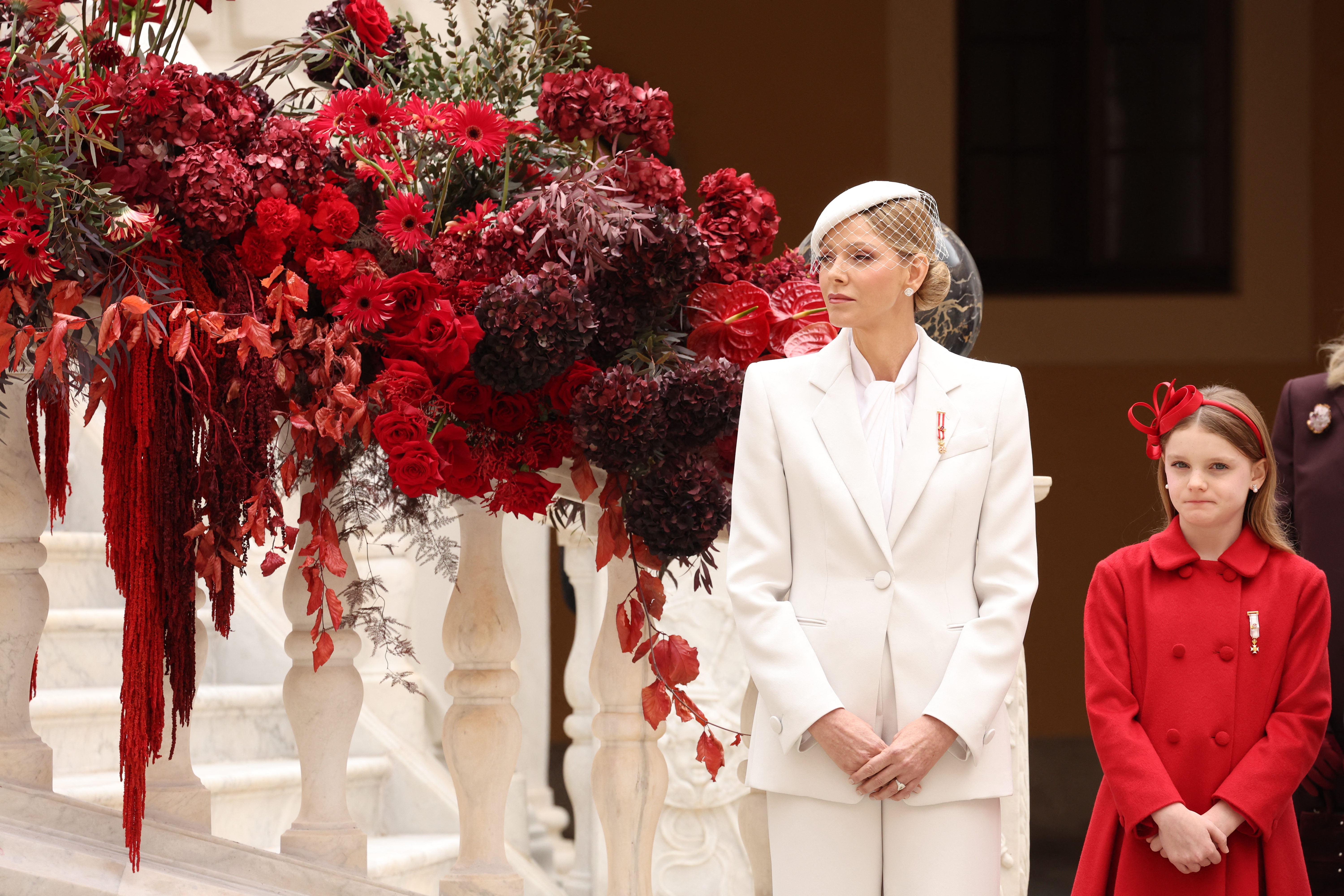Princess Charlene in a white suit and Princess Gabriella in a red coat dress