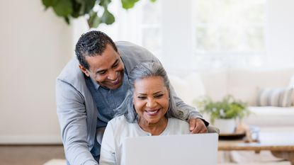 Smiling retirees look at a laptop together at their dining room table.