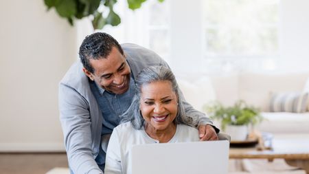 Smiling retirees look at a laptop together at their dining room table.
