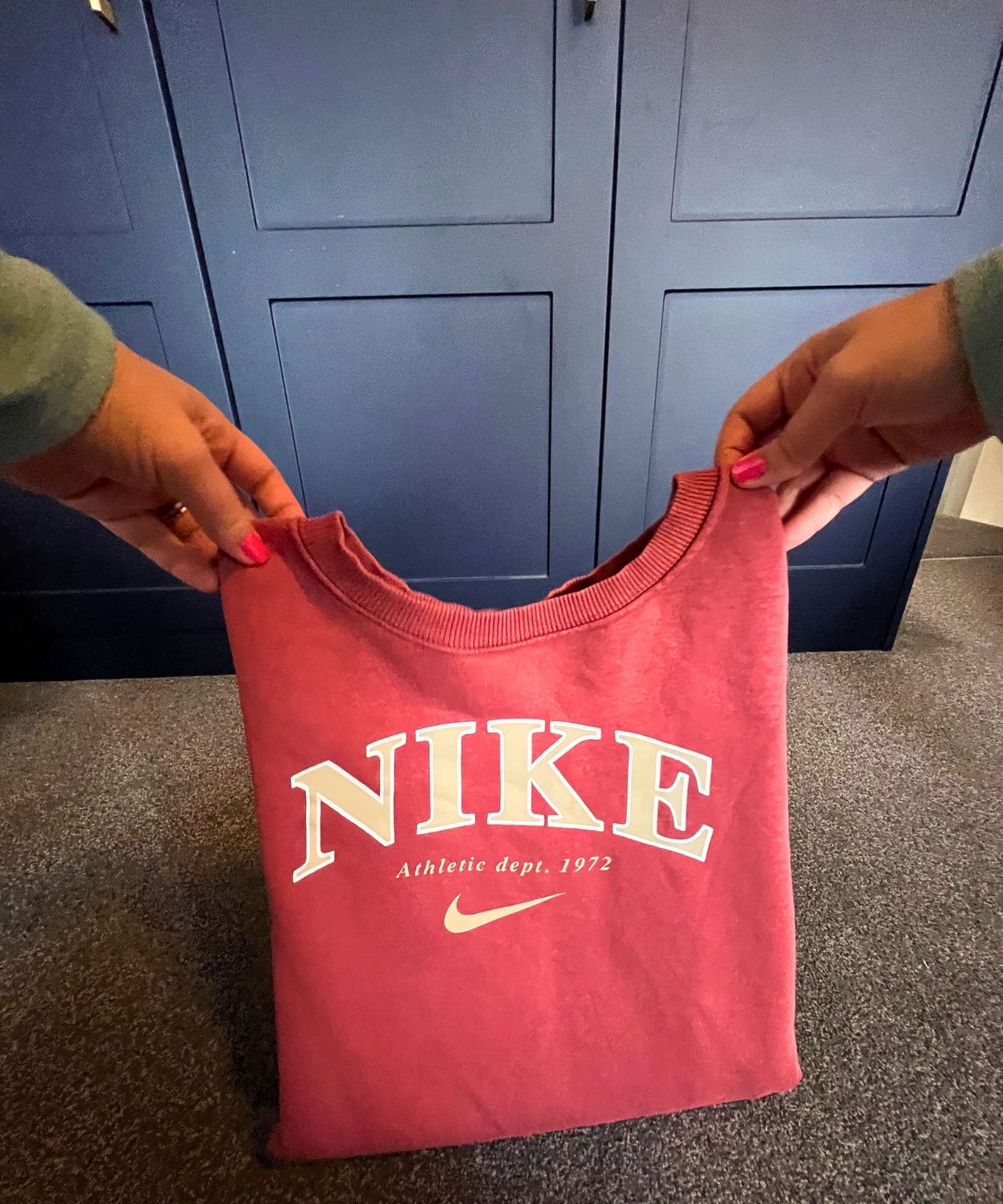 The image shows a point of view shot of a peachy-orange Nike long sleeve sweater being folded back over its bottom half on a gray carpeted floor