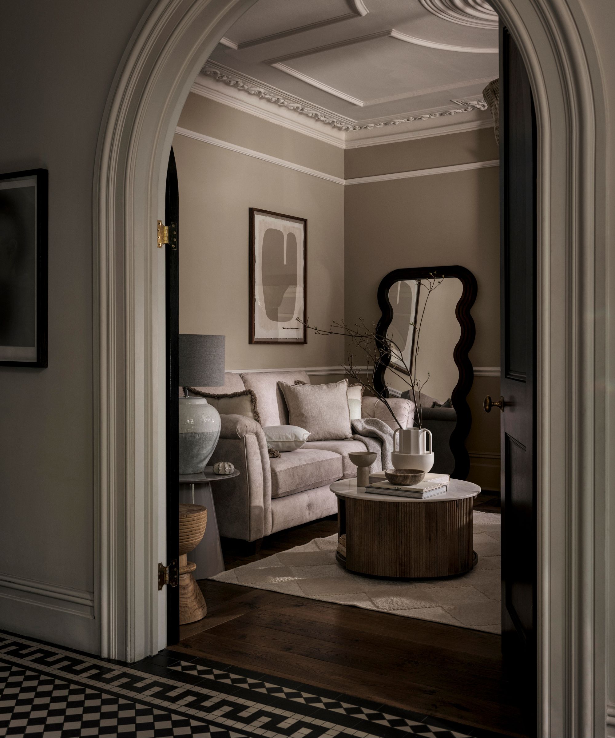 greige traditional victorian living room with dark wooden floors, a greige couch, round marble and wood coffee table, wavy floor length mirror and beige rug