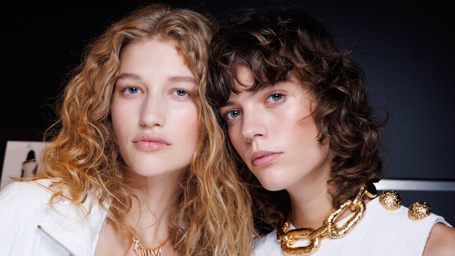 two models backstage at roberto cavalli spring 2025 fashion show wearing white shirts and gold necklaces with clean makeup 