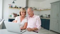 A retired couple sits at a table, applying for Social Security benefits on a laptop computer.